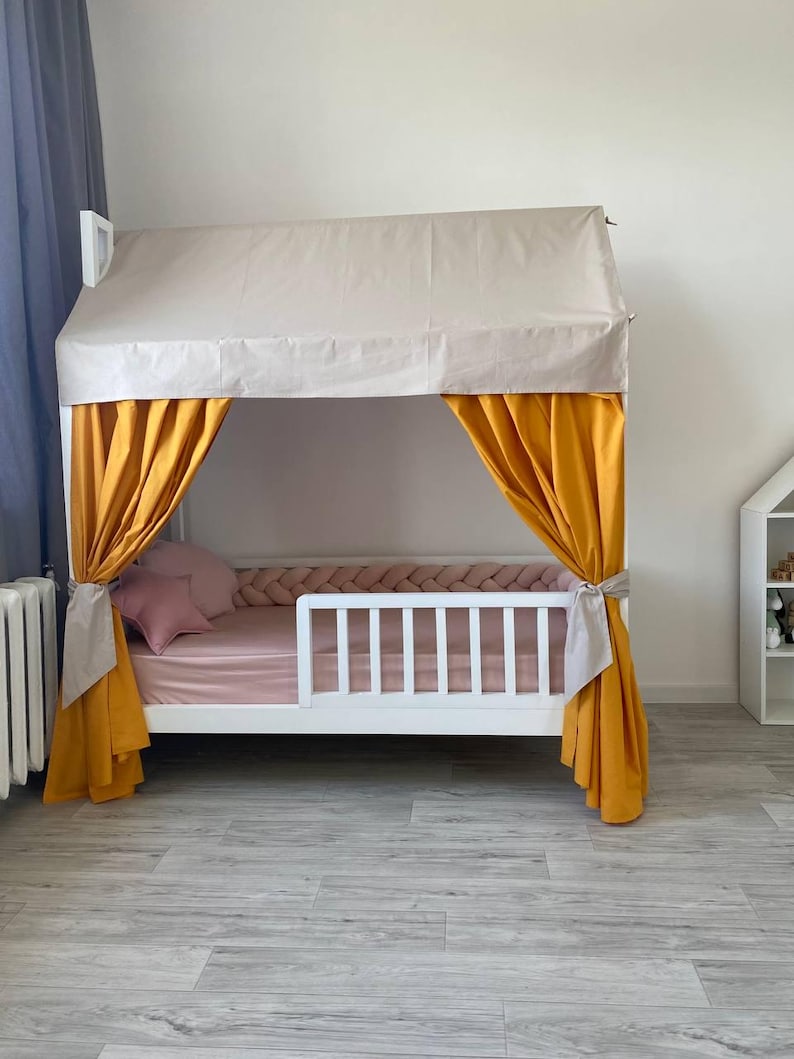 May include: A white wooden house-shaped bed frame featuring a beige fabric roof canopy, mustard yellow side curtains tied back, and a pink mattress with a matching pink star-shaped pillow and a braided pink bumper guard along the interior edge.
