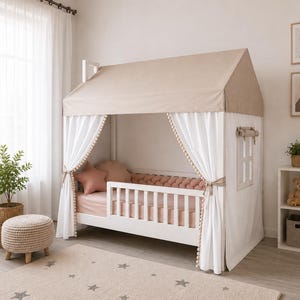 May include: A white wooden house-shaped bed frame featuring a beige fabric roof, white curtains with pom-pom trim, and a slatted side rail. The bed is dressed with pink sheets, a pink braided bumper, and star-shaped pillows in a soft pink hue.