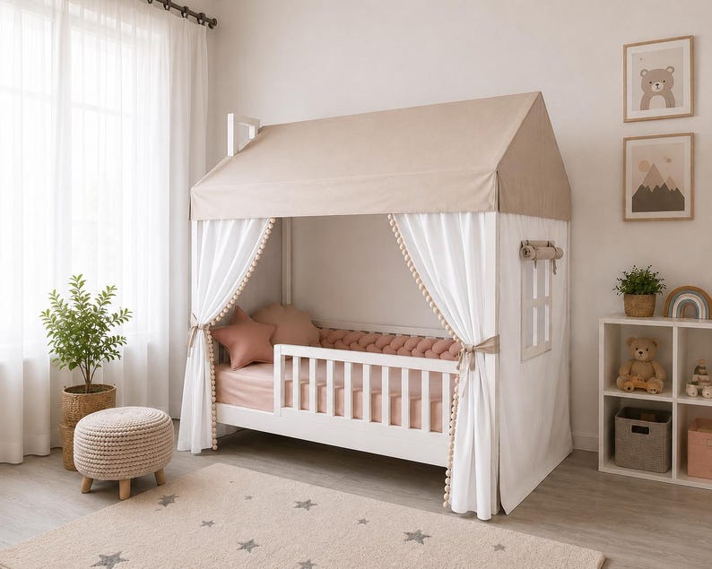 May include: A white wooden house-shaped bed frame featuring a beige fabric roof, white curtains with pom-pom trim, and a slatted side rail. The bed is dressed with pink sheets, a pink braided bumper, and star-shaped pillows in a soft pink hue.