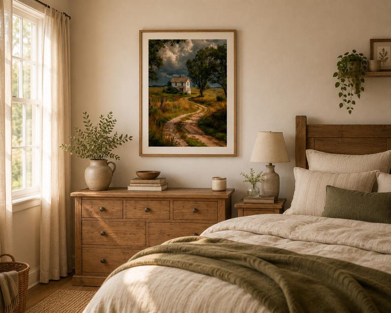 May include: A framed wall art print featuring a landscape painting of a white house at the end of a winding dirt path, surrounded by golden fields and dark, stormy clouds, displayed in a wooden frame on a light-colored wall above a dresser.