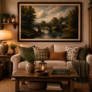 May include: A living room featuring a beige sofa with assorted throw pillows, a wooden coffee table with books and a lantern, and a large framed landscape painting of a stone bridge over a river hanging on the wall above the sofa.