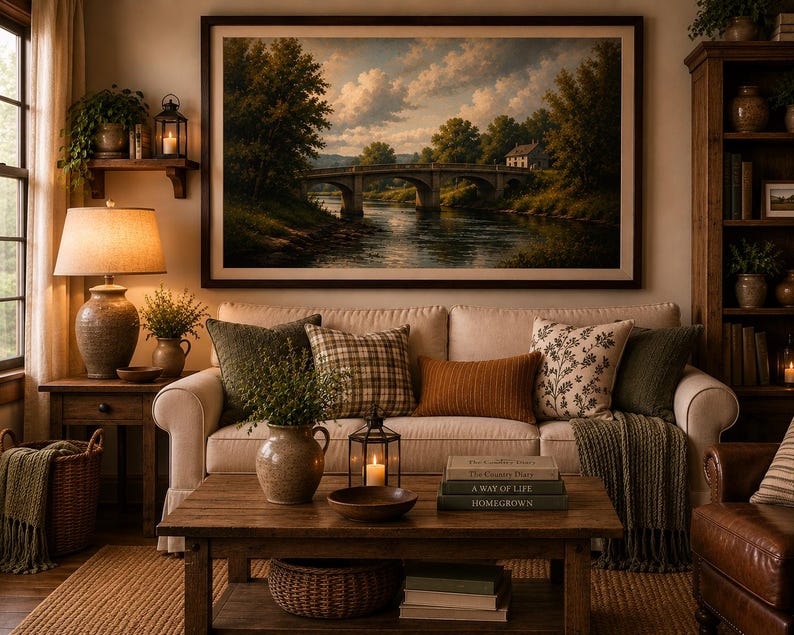 May include: A living room featuring a beige sofa with assorted throw pillows, a wooden coffee table with books and a lantern, and a large framed landscape painting of a stone bridge over a river hanging on the wall above the sofa.