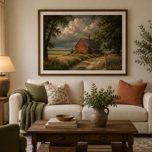 May include: A framed landscape wall art print featuring a red barn situated in a grassy field under a cloudy sky, displayed above a cream-coloured sofa with green and orange throw pillows, a wooden coffee table, and a ceramic table lamp.