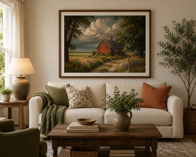 May include: A framed landscape wall art print featuring a red barn situated in a grassy field under a cloudy sky, displayed above a cream-coloured sofa with green and orange throw pillows, a wooden coffee table, and a ceramic table lamp.