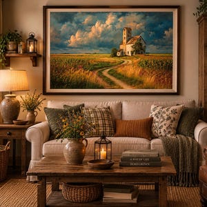 May include: A large framed landscape wall art print depicting a rural scene with a white building, a dirt path, and a field under a cloudy sky, displayed above a beige sofa with various throw pillows and a wooden coffee table in a living room.