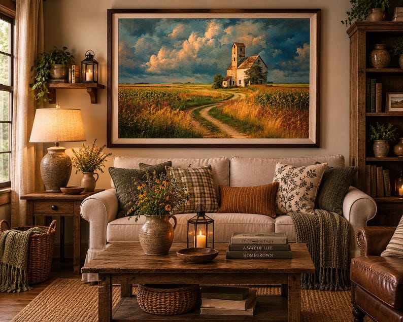 May include: A large framed landscape wall art print depicting a rural scene with a white building, a dirt path, and a field under a cloudy sky, displayed above a beige sofa with various throw pillows and a wooden coffee table in a living room.