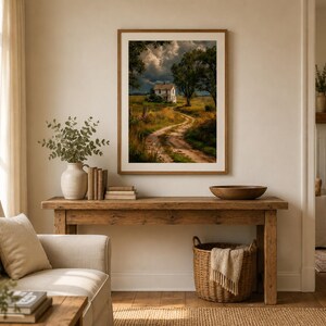 May include: A framed landscape print featuring a white house at the end of a winding dirt path through a grassy field under a dramatic, cloudy sky. The artwork is displayed in a light wood frame hanging on a neutral wall above a wooden console table.