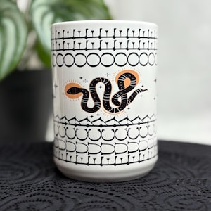 May include: White ceramic mug with a black and orange snake design. The mug features a repeating black pattern at the top and bottom, with a stylized snake in the center. The mug is cylindrical and sits on a black surface.
