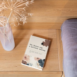 May include: A hardcover journal titled "The Thrive Log: a wellness journey" with floral illustrations in shades of brown and blue. The journal rests on a wooden surface next to a glass vase with dried flowers. A bed with a gray comforter is in the background.