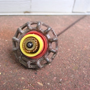 May include: Close-up of a vintage faucet handle. The handle is made of aged metal with a circular design. It features red, yellow, and brass-colored internal components. The handle is on a red brick surface.