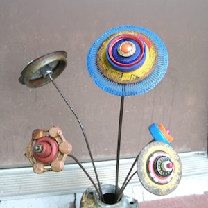 May include: A sculpture of four flowers made from repurposed metal and plastic objects. The flowers feature components like a blue plastic gear, a red valve handle, a metal lid, and a circular disc with the text LEGENDARY, all attached to thin metal stems.