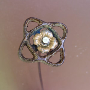 May include: An antique hat pin with a decorative head. The head features a gold-colored flower in the center, surrounded by a blue accent and an openwork design in a bronze-like metal. The pin is slender and appears to be made of metal.