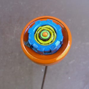 May include: A colorful, abstract pin with an orange, circular base and a central design of blue, yellow, and green plastic components. The pin has a thin, metal stem.