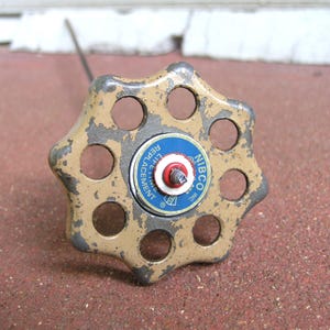 May include: A vintage, star-shaped valve handle with a worn, tan and gray finish. The center features a blue and white label with the text "NIBCO LIFE REPLACEMENT". The handle has multiple circular openings.