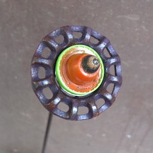 May include: A handmade garden stake featuring a repurposed metal wheel with a rusty, dark purple finish. The center has a bright orange and green design. The stake is perfect for adding a unique touch to any garden or outdoor space.