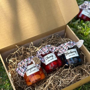 May include: A gift box containing three jam candles in glass jars. The jars are labeled "Tangerine", "Cherry", and "Blueberry". Each jar is topped with a plaid fabric lid and a white label. The box is filled with shredded paper.