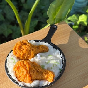 May include: A candle shaped like a skillet of fried chicken and mashed potatoes. The candle features two orange fried chicken drumsticks atop a bed of white mashed potatoes, with green sprinkles. The skillet is black and sits on a wooden surface.