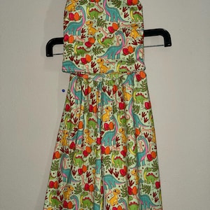 May include: A two-piece outfit featuring a halter top and wide-leg pants. The set is light green with a repeating pattern of colorful dinosaurs, eggs, and foliage. The dinosaurs are in shades of blue, green, and yellow.