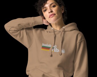 May include: A tan hoodie with a front pocket and a drawstring hood. The hoodie features the text "Isaiah 48 Vibes" in a colorful, retro-style design. The hoodie is made of a soft material and is suitable for casual wear.