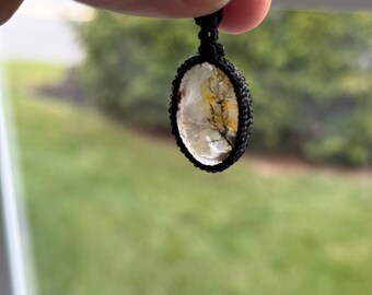May include: Oval pendant with a black cord. The pendant features a clear stone with yellow and brown inclusions, encased in a black woven setting. The background is a blurred view of green foliage.