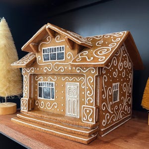 Puede incluir: Una casa de jengibre con detalles de glaseado blanco y dos pequeños árboles decorativos. La casa tiene un techo detallado, ventanas y una puerta. Los árboles son amarillos y naranjas, añadiendo un toque festivo.