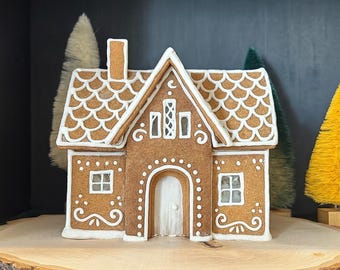 May include: A gingerbread house with white icing details, including roof shingles, window frames, and door trim. The house has a chimney and is decorated with swirls and dots. The house is displayed on a wooden slice.