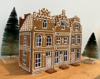 May include: A gingerbread house display featuring three brown houses with white icing details. Each house has windows, doors, and decorative trim. Two small green Christmas trees flank the houses, creating a festive holiday scene.