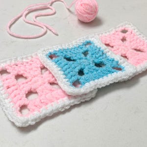 May include: Handmade crochet squares in pink and blue with white edging. A ball of pink yarn and loose yarn strands are in the background. The squares are a classic granny square design, ideal for crafting projects.