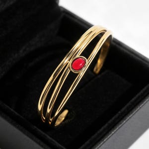 Puede incluir: Un brazalete dorado con tres bandas paralelas que convergen en un punto central. El centro del brazalete está adornado con una única piedra roja lisa de forma ovalada, engastada en un bisel dorado, presentado en una caja de color negro.