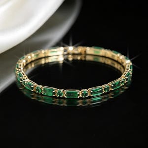 May include: A gold bangle bracelet featuring a continuous row of alternating rectangular and round green gemstones. The metal setting is polished gold, and the stones are set in a repeating pattern around the entire circumference of the circular band.