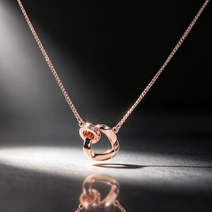 May include: A rose gold-toned necklace featuring a delicate chain and a pendant composed of two interlocking, smooth, circular metal rings. The design is polished and reflective, catching light to create a subtle glow on the metallic surfaces.