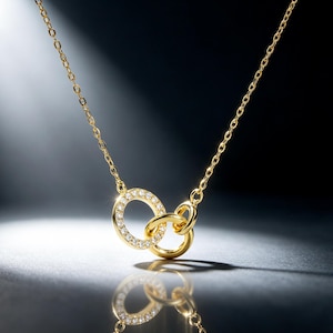 May include: A gold-toned necklace featuring two interlocking circular pendants. One circle is embellished with a series of small, clear, sparkling stones along its outer edge, while the second circle is a plain, polished gold-toned metal ring.