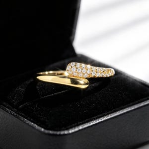 May include: A gold bypass ring featuring a polished band on one side and a curved end encrusted with small, clear, sparkling stones. The ring is presented inside a black, velvet-lined jewelry box with a hinged lid, showcasing a reflective finish.