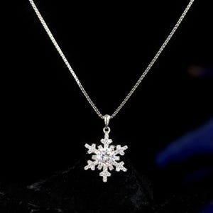 Op de afbeelding: Een zilveren ketting met een sneeuwvlok hanger. De hanger is een gedetailleerd sneeuwvlokontwerp met een grote, heldere middensteen en kleinere stenen op elk punt. De ketting is delicaat en hangt tegen een donkere achtergrond.