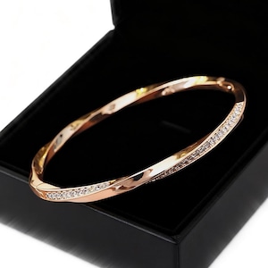 May include: A rose gold bangle bracelet with a twisted design, set with small, sparkling clear stones along one side. The bracelet is displayed inside a black jewelry box, creating a contrast that highlights the bracelet's elegance. A beautiful piece of jewelry.