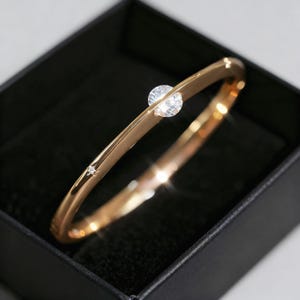 May include: A gold-colored bangle bracelet with a large, round, clear gemstone and a smaller, clear gemstone. The bracelet is displayed in a black box. The bracelet has a smooth, polished finish and a simple, elegant design.