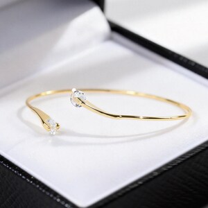 May include: A gold-toned open cuff bracelet featuring a thin, polished metal band. The two ends of the bracelet are slightly tapered and each is set with a single, clear, round-cut gemstone, creating a delicate and symmetrical design.