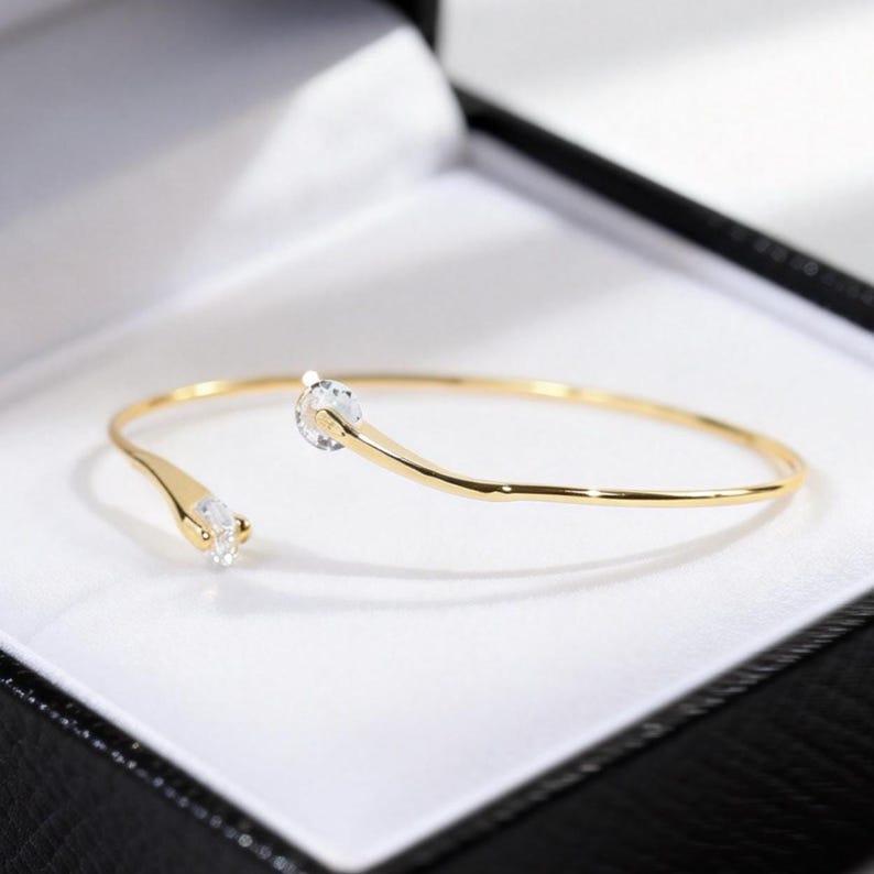 May include: A gold-toned open cuff bracelet featuring a thin, polished metal band. The two ends of the bracelet are slightly tapered and each is set with a single, clear, round-cut gemstone, creating a delicate and symmetrical design.