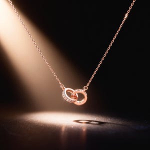 May include: Rose gold necklace with two intertwined rings, one set with small, clear gemstones. The fine chain and rings are displayed against a dark backdrop, lit by a gentle light.