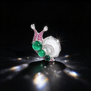 May include: A snail-shaped brooch crafted from silver-toned metal, featuring a white shell, three round green cabochon stones along the body, and small pink and clear crystals embedded throughout the head and shell structure.