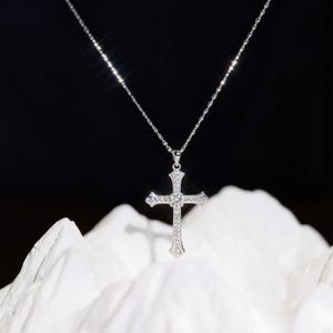 May include: A silver cross pendant necklace with a delicate chain. The cross is adorned with small, sparkling stones, and hangs against a dark background. The necklace is displayed on a white, textured surface.