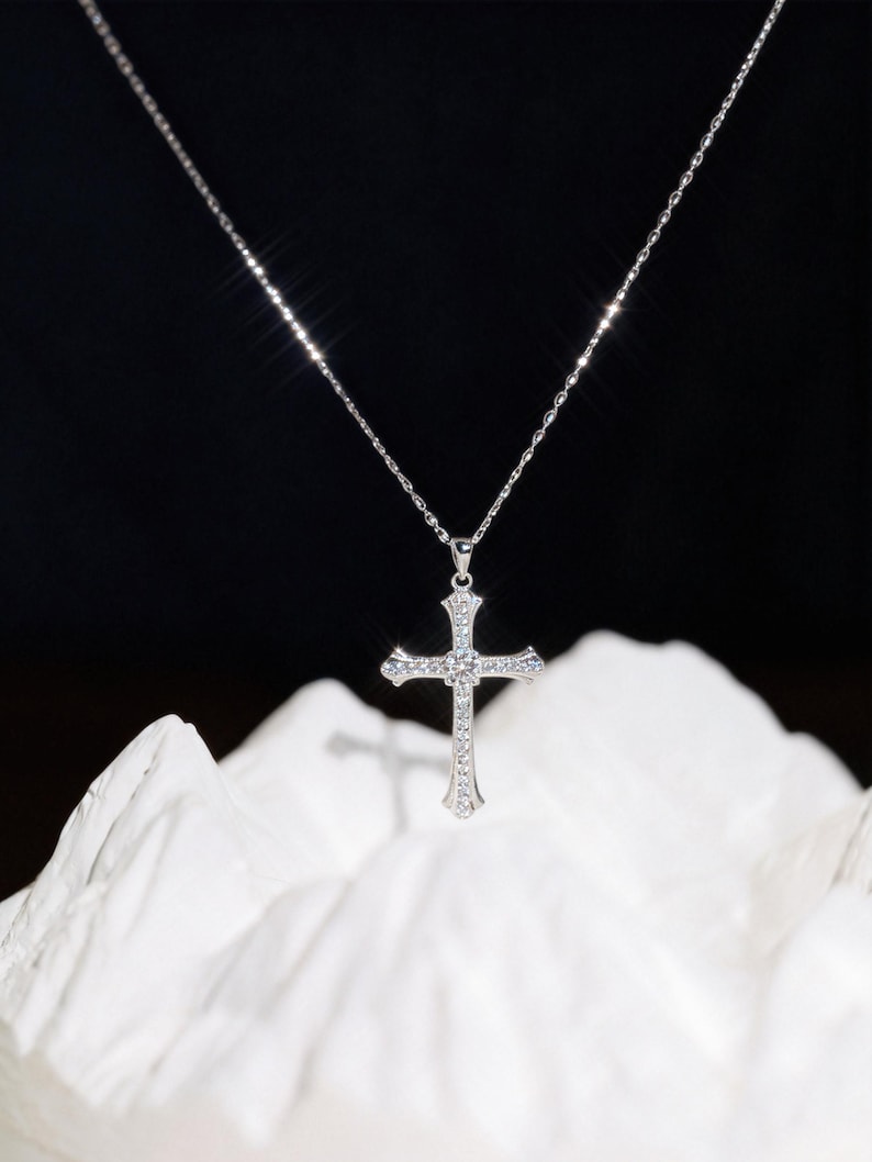 May include: A silver cross pendant necklace with a delicate chain. The cross is adorned with small, sparkling stones, and hangs against a dark background. The necklace is displayed on a white, textured surface.