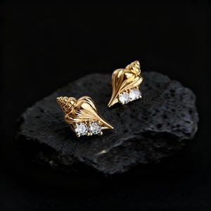 May include: A pair of gold-colored seashell stud earrings. Each earring features a detailed shell design with two clear, round gemstones. The earrings are displayed on a dark, textured surface, creating a contrast with the shiny gold.