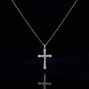 May include: A silver-colored cross pendant necklace with a delicate chain. The cross is adorned with small, sparkling gemstones, and is displayed against a black background. The necklace is reflected in the glossy surface below.
