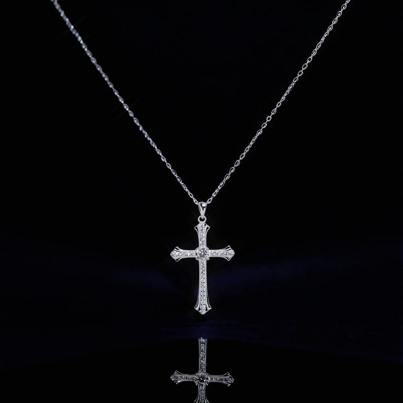 May include: A silver-colored cross pendant necklace with a delicate chain. The cross is adorned with small, sparkling gemstones, and is displayed against a black background. The necklace is reflected in the glossy surface below.