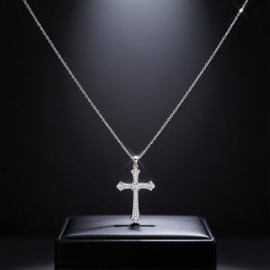 May include: A silver-colored cross pendant necklace with a delicate chain. The cross is adorned with small, sparkling gemstones. The necklace is displayed on a black surface against a dark background.
