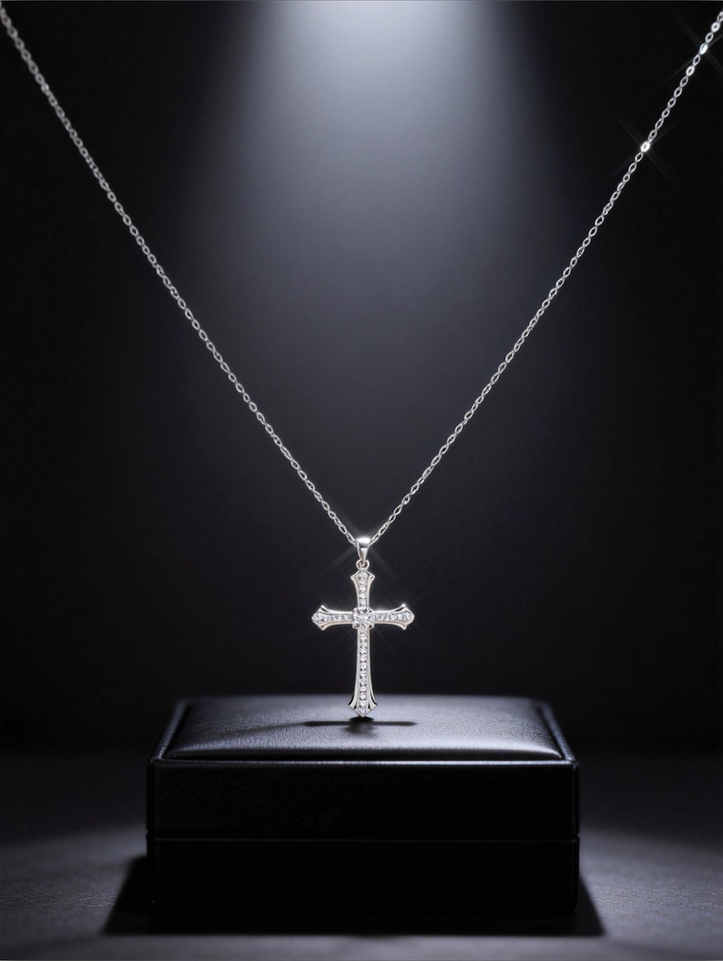 May include: A silver-colored cross pendant necklace with a delicate chain. The cross is adorned with small, sparkling gemstones. The necklace is displayed on a black surface against a dark background.