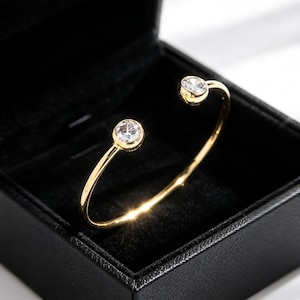 May include: A gold-toned open cuff bracelet featuring two round, clear, faceted stones set at each end. The bracelet has a smooth, polished finish and is displayed inside a black, square-shaped jewellery box with a soft, dark interior lining.