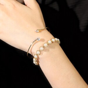 May include: A gold-toned open cuff bracelet featuring a single clear stone at each end, paired with a gold-toned chain bracelet composed of circular links, each set with a small white pearl, creating a layered look on the wrist.