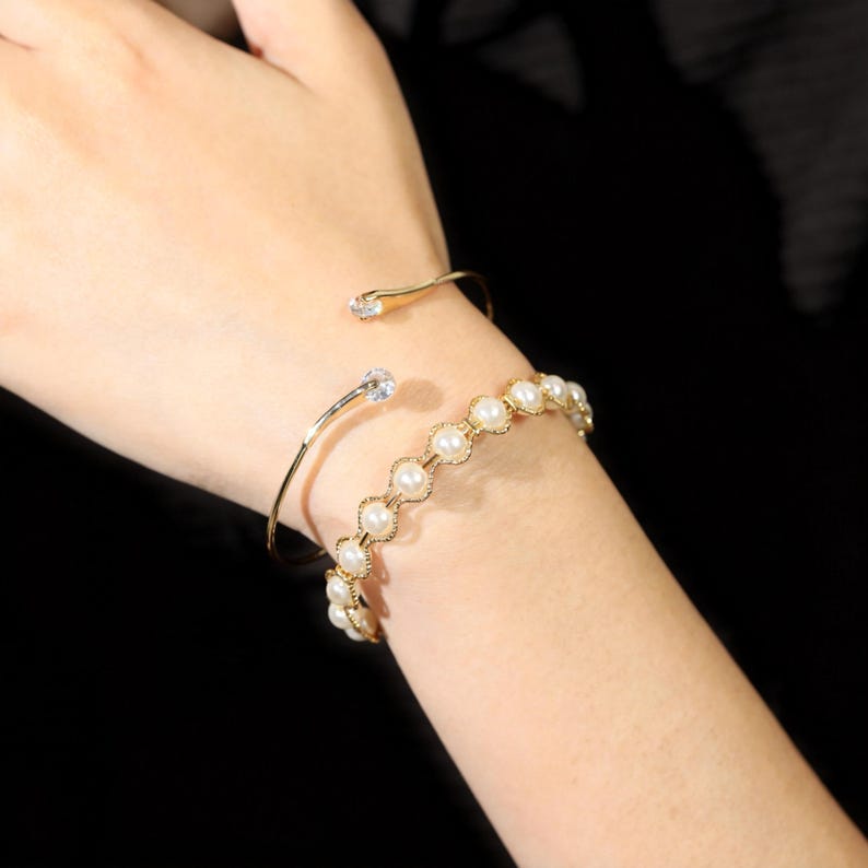 May include: A gold-toned open cuff bracelet featuring a single clear stone at each end, paired with a gold-toned chain bracelet composed of circular links, each set with a small white pearl, creating a layered look on the wrist.